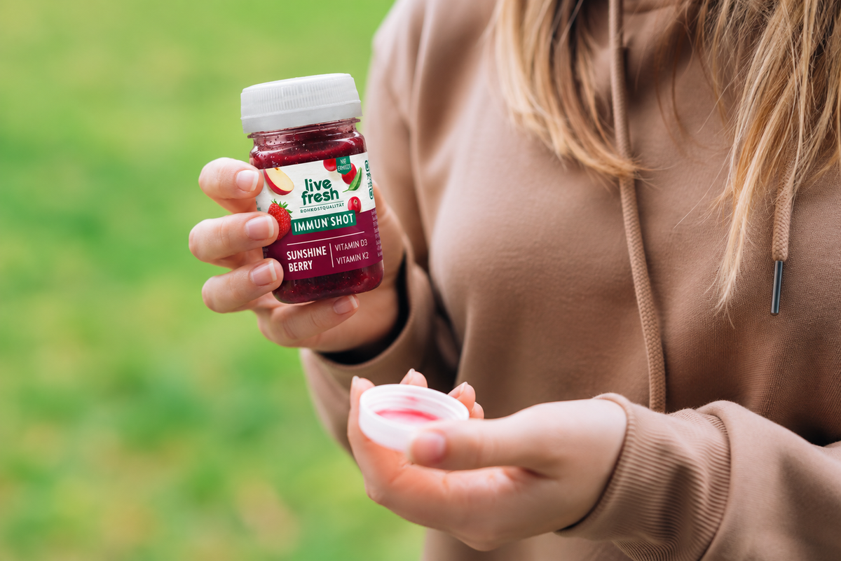 Eine Frau hält draußen den roten Live Fresh Immun Shot Sunshine Berry mit Vitamin D3 und Vitamin K2 in der Hand. Die neue kompakte Flaschenform mit weißem Schraubdeckel und dunkelrotem Shot steht vor einem unscharfen grünen Hintergrund im Fokus.