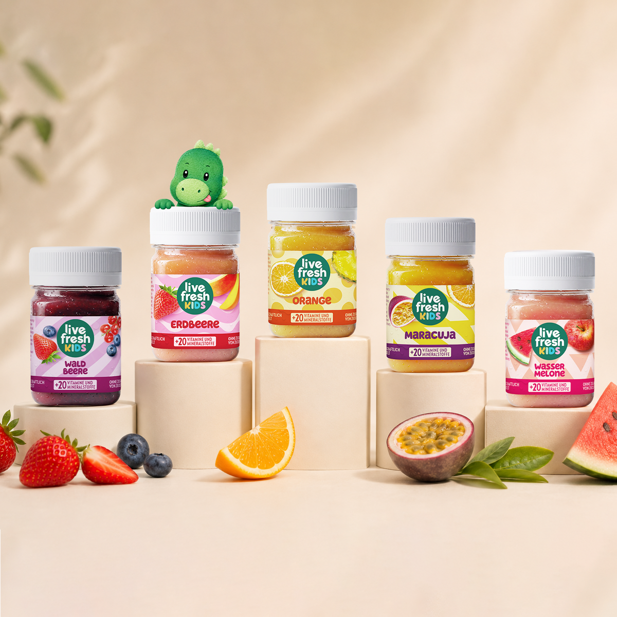 Five jars labeled “Live Fresh Kids,” “Wild Berries,” “Strawberry,” “Orange,” and “Passion Fruit.” "Watermelon" and "+20 Vitamins and Minerals" are placed on pedestals of varying heights. A small green dinosaur peeks out from behind the strawberry jar, and fresh fruit lies in front of it.