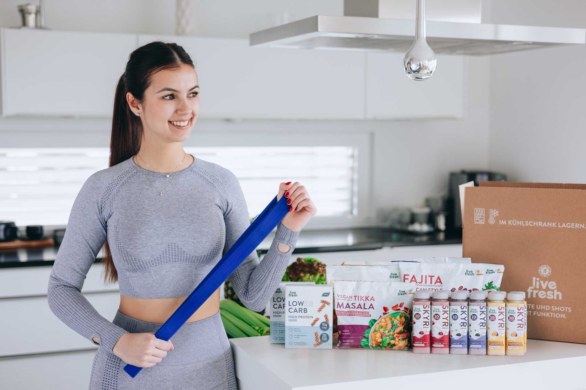 Frau in sportlicher Kleidung, die in einer modernen Küche steht und ein blaues Fitnessband hält. Auf der Kücheninsel vor ihr sind mehrere Produkte von „Live Fresh“ zu sehen, darunter verschiedene Getränke und Lebensmittel wie Low-Carb High-Protein-Nudeln, veganes Tikka Masala und Fajita-Mischungen. Neben den Produkten steht ein großer brauner Karton mit dem Aufdruck „Live Fresh“ und der Anweisung „Im Kühlschrank lagern“. Die Frau lächelt und wirkt bereit für ein Workout oder eine gesunde Mahlzeit.