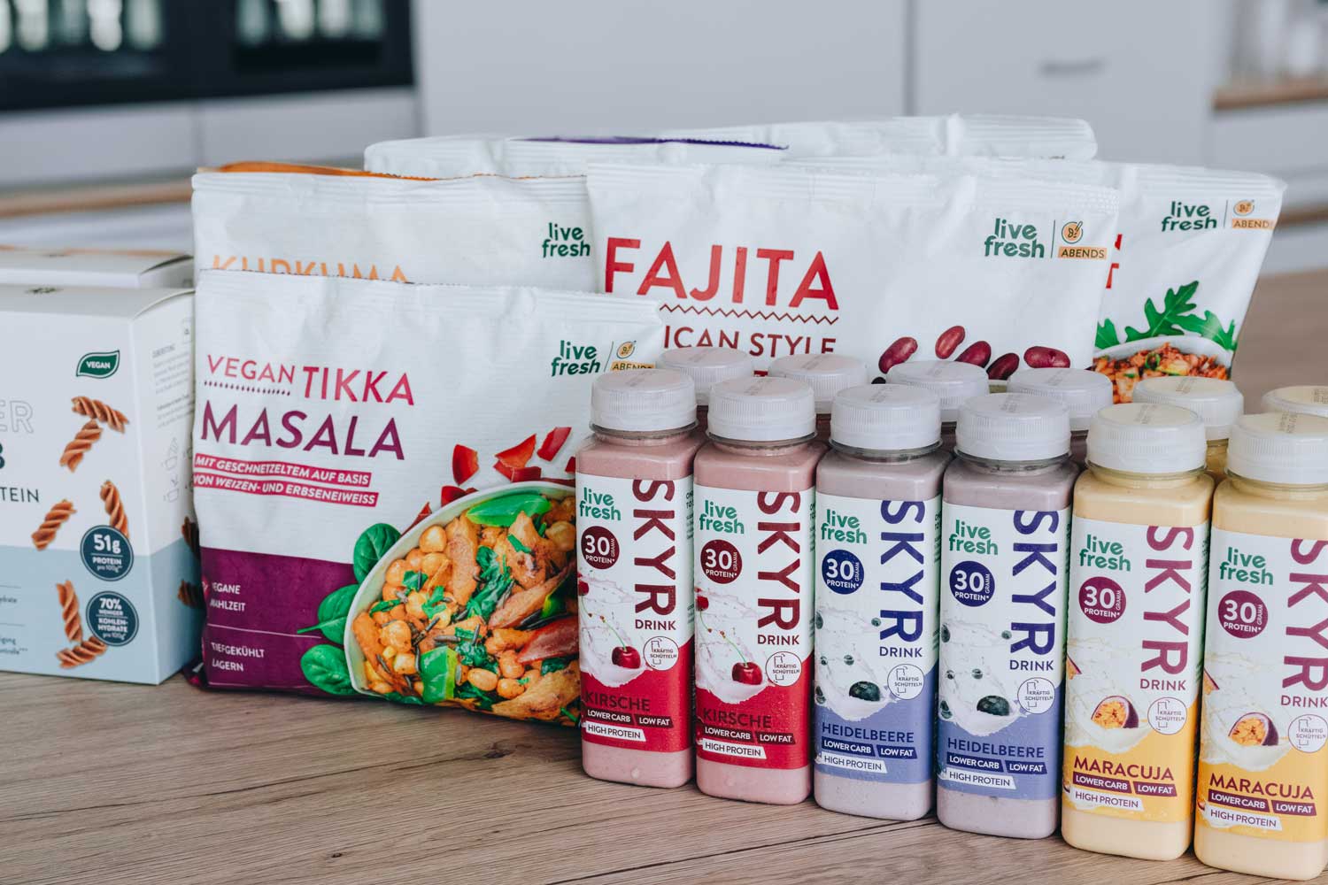 Auswahl von „Live Fresh“-Produkten, die auf einer Arbeitsplatte präsentiert werden. Zu sehen sind verschiedene gefrorene Mahlzeiten wie „Vegan Tikka Masala“ und „Fajita Mexican Style“. Daneben stehen mehrere Flaschen „Skyr Drink“ in verschiedenen Geschmacksrichtungen, darunter Kirsche, Heidelbeere und Maracuja. Auf der linken Seite des Bildes ist eine Packung Low-Carb High-Protein-Nudeln zu sehen. Alle Produkte sind als proteinreich und für eine gesunde Ernährung geeignet gekennzeichnet.