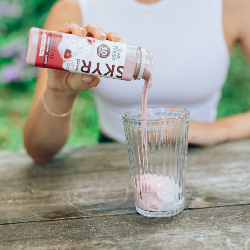 Eine Person, die „Live Fresh Skyr Drink Kirsche“ in ein Glas gießt. Der rosa Skyr-Drink, der 30g Protein enthält, wird betont als proteinreich, fettarm und kohlenhydratreduziert. Die Person trägt ein weißes Oberteil, und die Szene spielt sich draußen an einem Holztisch ab, was eine entspannte und gesunde Atmosphäre suggeriert.