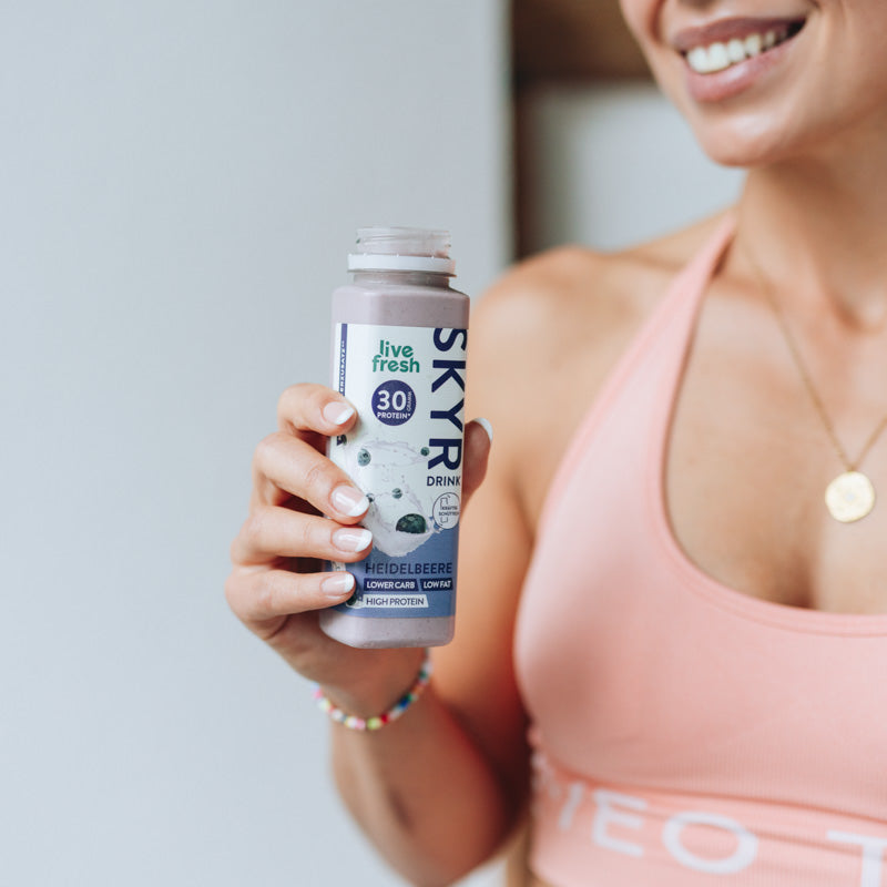 Eine Frau in Sportbekleidung, die eine offene Flasche „Live Fresh Skyr Drink Heidelbeere“ in der Hand hält. Die Skyr-Flasche hebt 30g Protein, niedrigen Kohlenhydratgehalt, fettarm und proteinreich hervor. Die Frau lächelt und trägt ein rosa Sportoberteil, was auf eine gesunde und aktive Lebensweise hindeutet.