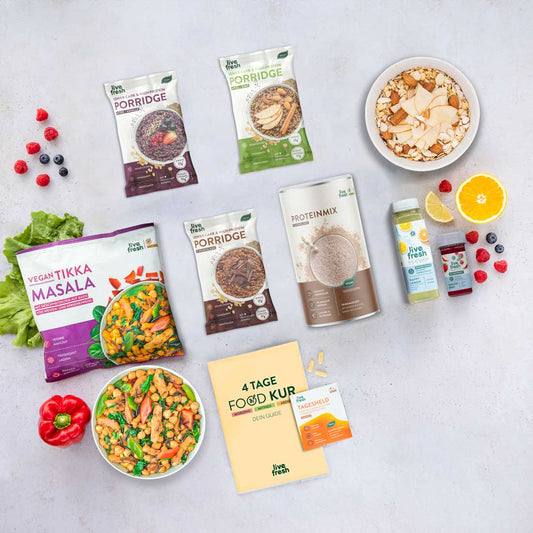 Flatlay mit verschiedenen Produkten aus der Tage-Foodkur/Abnehmbundles von LiveFresh: Porridges, Proteinmix, Tikka Masala, Säfte, Shots, ein Foodkur-Guide und Vitaminpräparate. Umgeben von frischem Obst und Gemüse.