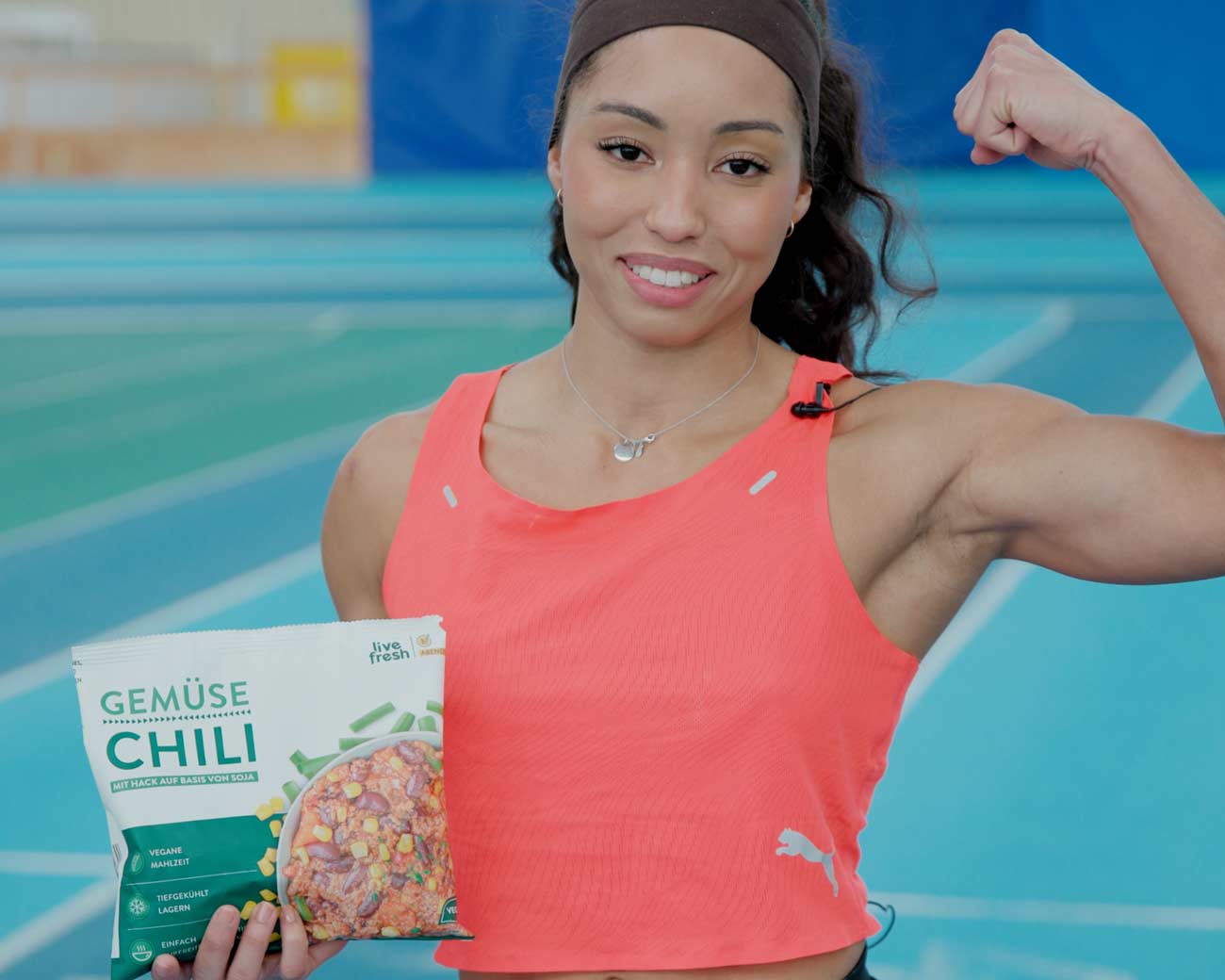 Eine Sportlerin in einem leuchtend orangefarbenen Top, die stolz ihre Arm-Muskeln zeigt. In ihrer anderen Hand hält sie eine Packung "Gemüse Chili" von Live Fresh, eine vegane Tiefkühlmahlzeit auf Sojabasis. Im Hintergrund ist eine blaue Sporthalle zu sehen.