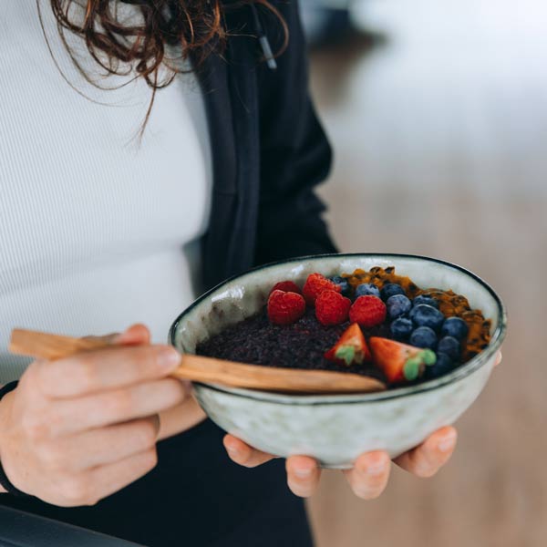 Eine Person, die eine Schüssel mit Porridge hält, garniert mit frischen Beeren wie Himbeeren, Heidelbeeren und Erdbeeren sowie weiteren Zutaten. Die Person isst den Porridge mit einem Holzlöffel und trägt sportliche Kleidung, wobei der Fokus auf der Schüssel liegt.