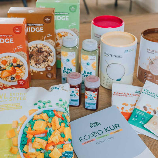 Auswahl von „Live Fresh“ Produkten, die auf einem Tisch angeordnet sind. Zu sehen sind mehrere Porridge-Packungen in verschiedenen Geschmacksrichtungen, Flaschen mit Säften, Proteinmischungen in Dosen sowie Snacks wie der „Veggie Mix“ und „Tamari Soja“. Zudem liegt ein „Food Kur“-Guide auf dem Tisch, der auf eine strukturierte Ernährungsplanung hinweist. Die Vielfalt der Produkte deutet auf eine komplette Kur oder Ernährungsumstellung hin, die sowohl flüssige als auch feste Mahlzeiten umfasst.