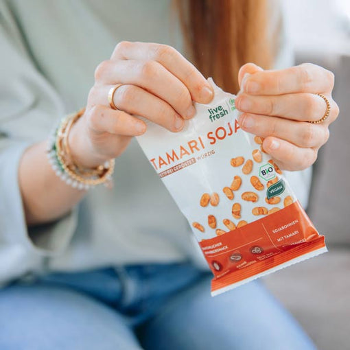 Zeigt eine Person, die eine Packung „Live Fresh Tamari Soja“-Snacks öffnet. Die Packung weist darauf hin, dass die Sojabohnen biologisch, vegan und mit Tamari gewürzt sind. Der Snack wird als gesunde, proteinreiche Option präsentiert. Die Szene vermittelt einen Moment der Entspannung und bewussten Ernährung.