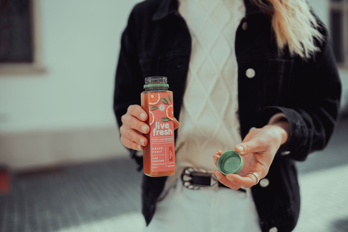 Eine Person, die eine geöffnete Flasche LiveFresh Grapefruit-Saft in der einen Hand und den grünen Deckel der Flasche in der anderen hält. Das Etikett auf der Flasche beschreibt den Saft als „Frisch & Kaltgepresst, Keine Zusätze – Nie Erhitzt“ sowie „100% Frischer Grapefruit Saft“. Die Person trägt eine weiße Strickjacke unter einer schwarzen Jacke und steht in einer hellen Umgebung.