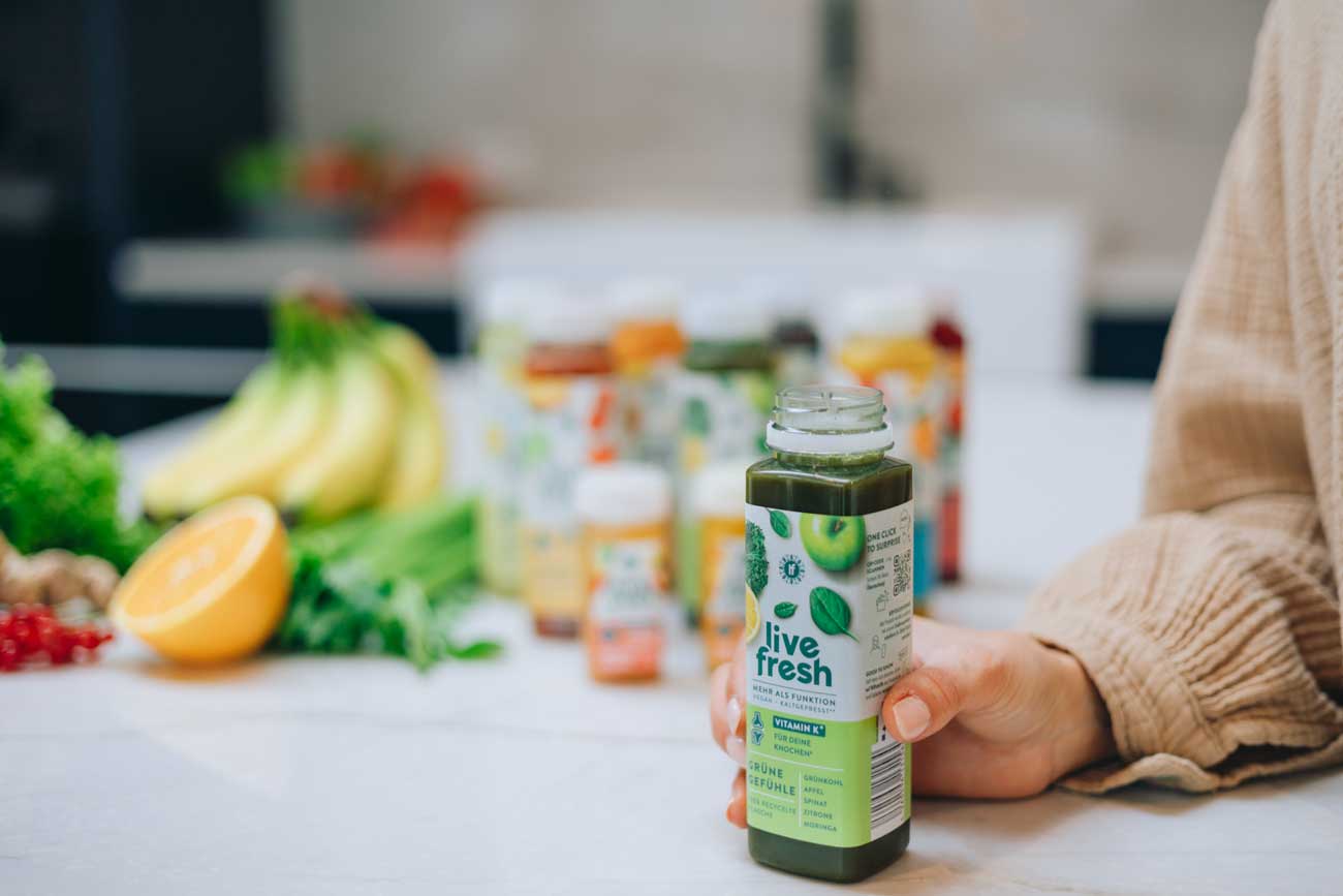 Eine Nahaufnahme einer Hand, die eine Flasche „Live Fresh“ Saft hält. Auf der Flasche steht „Grüne Gefühle“, ein Saft mit Zutaten wie Grünkohl, Apfel, Limette, Zitrone und Moringa. Im Hintergrund sind verschiedene Obst- und Gemüsearten, darunter Bananen und eine halbe Orange, sowie weitere „Live Fresh“-Produkte zu sehen. Die Szene vermittelt Frische und gesunde Ernährung.