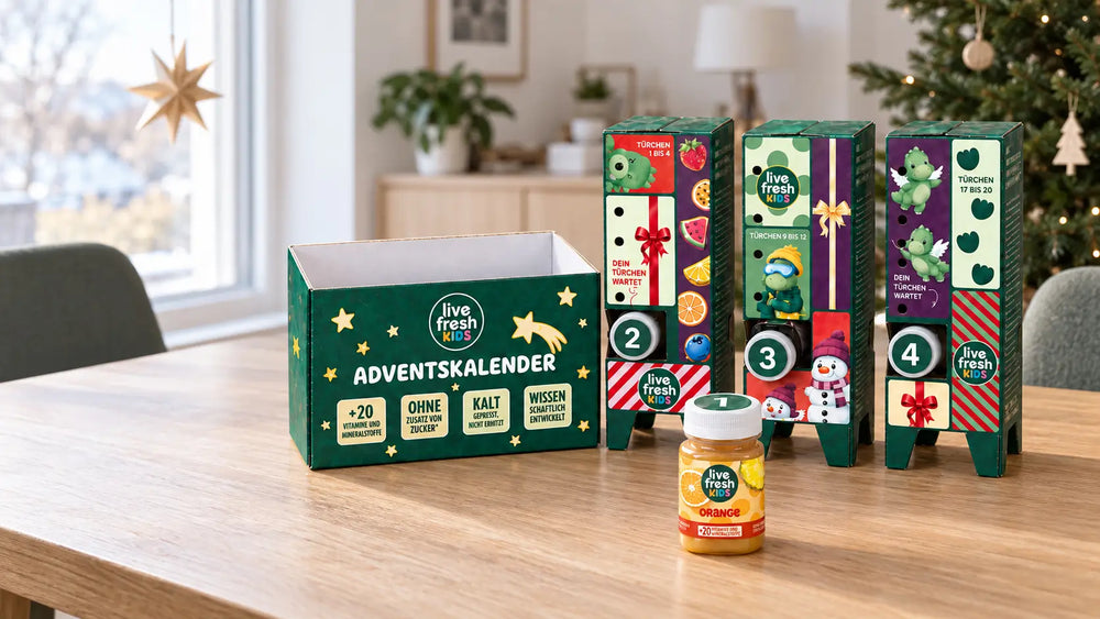 Der geöffnete LiveFresh Kids Adventskalender steht auf einem Holztisch vor einem geschmückten Weihnachtsbaum mit der Flasche Orange aus Türchen eins davor. Auf der Verpackung steht "ADVENTSKALENDER" sowie "+20 VITAMINE UND MINERALSTOFFE" "OHNE ZUSATZ VON ZUCKER" "KALT GEPRESST NICHT ERHITZT" und "WISSENSCHAFTLICH ENTWICKELT".