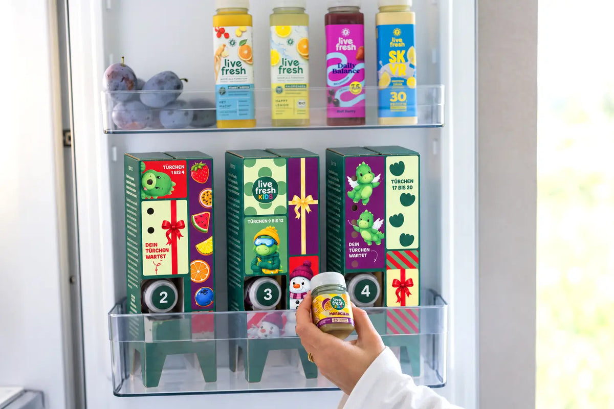 Eine Hand nimmt eine Flasche LiveFresh Kids Maracuja aus einem Kühlschrank. In der Kühlschranktür stehen der farbenfrohe Adventskalender sowie weitere LiveFresh Getränke und frisches Obst.