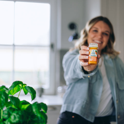 Eine Frau, die lächelnd im Hintergrund einen Kurkuma Power Shot von LiveFresh in die Kamera hält. 