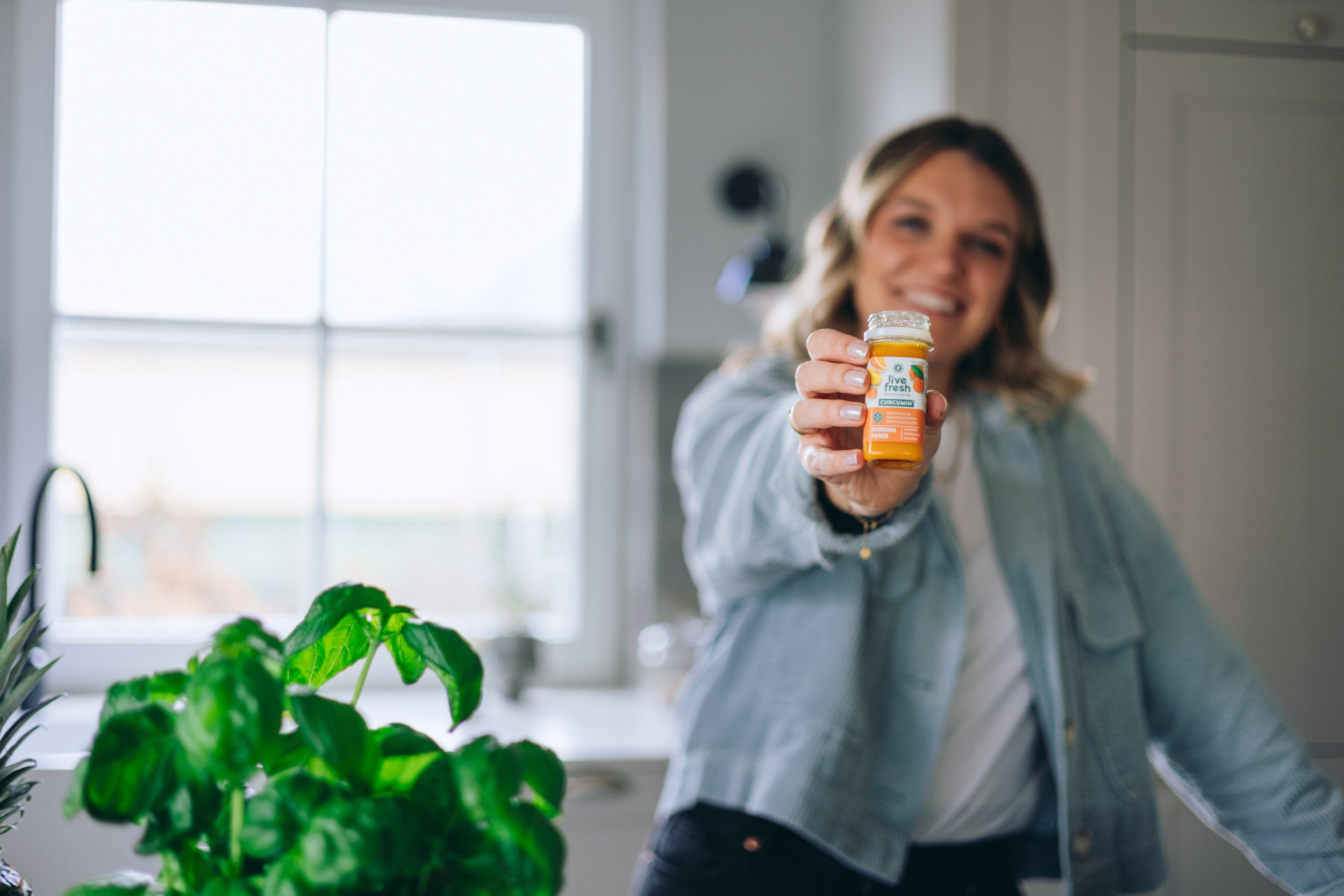 Eine Frau, die lächelnd im Hintergrund einen Kurkuma Power Shot von LiveFresh in die Kamera hält. 