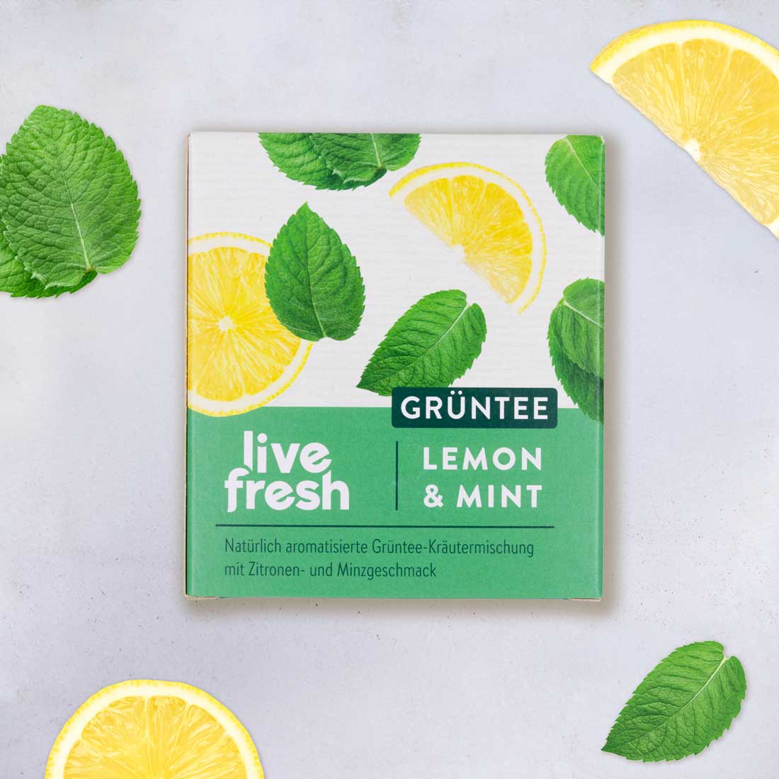 Verpackung des LiveFresh Grüntees „Lemon & Mint“ liegt auf hellem Untergrund, umgeben von frischen Zitronenscheiben und Minzblättern.