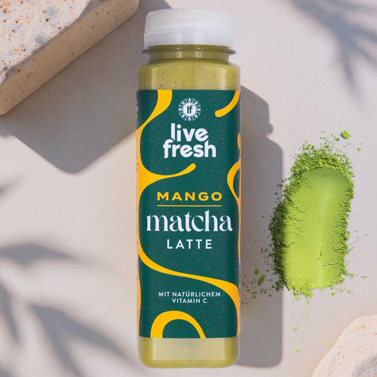 „Live Fresh“ Mango Matcha Latte Flasche neben Matcha-Pulver, arrangiert auf hellem Untergrund mit natürlichem Schattenwurf.