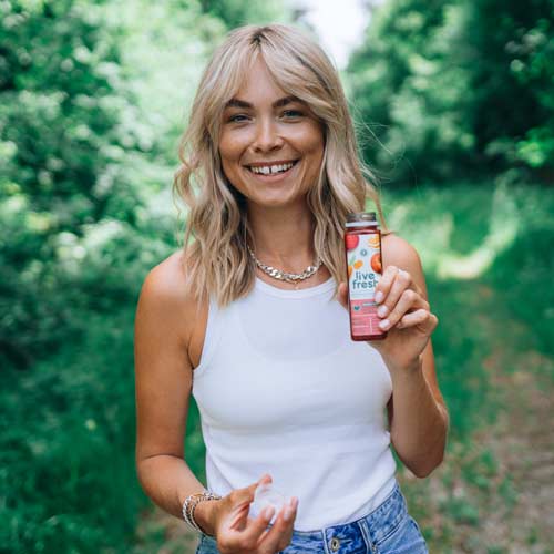 Eine blonde Frau, die draußen auf einem grasbewachsenen Weg steht und eine Flasche Saft in der Hand hält. Auf der Flasche steht "Live Fresh" und es sind Früchte abgebildet, was auf einen frischen Saft oder Smoothie hinweist. Die Frau trägt ein weißes ärmelloses Oberteil und lächelt fröhlich in die Kamera. Im Hintergrund ist eine grüne, bewaldete Umgebung zu sehen.