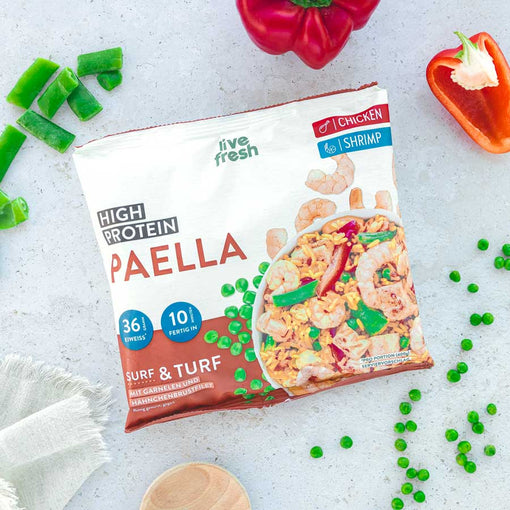 Das Bild zeigt eine Packung „Live Fresh High Protein Paella“ in der Surf & Turf-Variante mit Garnelen und Hähnchenbrustfilet. Die Verpackung hebt den hohen Proteingehalt von 36 g und eine Zubereitungszeit von 10 Minuten hervor. Um die Packung herum sind frische Zutaten wie grüne Paprika, Erbsen und rote Paprika verteilt, was die frische Zubereitung der Paella unterstreicht.