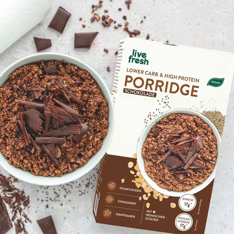Schoko-Porridge: Eine Schüssel mit Schokoladen-Porridge, garniert mit dunklen Schokoladenraspeln. Um die Schüssel herum sind Schokoladenstücke und Kakaosplitter verteilt. Im Hintergrund die Verpackung von "Live Fresh Lower Carb & High Protein Porridge Schokolade" mit brauner Akzentfarbe.