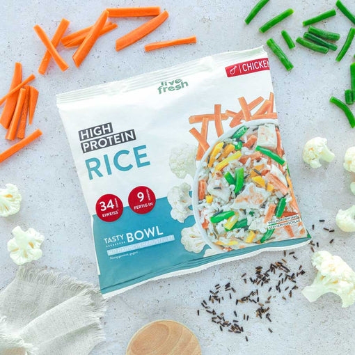 Das Bild zeigt eine Packung „Live Fresh High Protein Rice“ Tasty Bowl mit Hähnchenbrustfilet. Die Verpackung hebt einen Proteingehalt von 34 g und eine Zubereitungszeit von 9 Minuten hervor. Rund um die Packung liegen frische Zutaten wie Karotten, grüne Bohnen und Blumenkohl, was die ausgewogene und proteinreiche Mahlzeit betont.