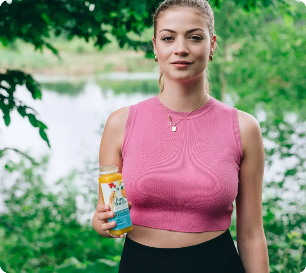 Blonde Frau in pinkem Top hält eine geöffnete Flasche Live Fresh-Saft, im Hintergrund grüner Seeuferbereich mit Bäumen.