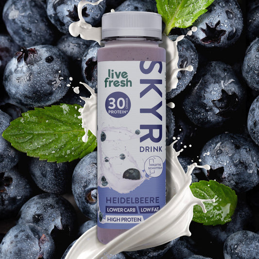 Nahaufnahme einer Flasche Skyr-Drink Heidelbeere von 'live fresh', umgeben von frischen Heidelbeeren und Minzblättern. Auf der Flasche steht '30g Protein', 'lower carb, low fat, high protein' und 'kräftig schütteln'. Weiße Spritzer suggerieren, dass der Drink cremig ist.