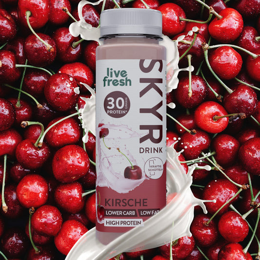 Nahaufnahme einer Flasche Skyr-Drink Kirsche von 'live fresh', umgeben von frischen, roten Kirschen. Auf der Flasche steht '30g Protein', 'lower carb, low fat, high protein' und 'kräftig schütteln'. Weiße Spritzer verdeutlichen die cremige Textur des Drinks.