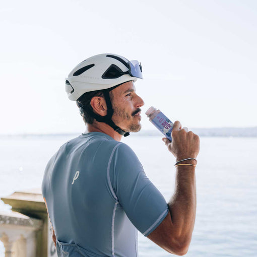 Radfahrer mit weißem Helm und blaugrauem Trikot trinkt ein Skyr-Drink von LiveFresh mit Blick aufs Wasser bei hellem Tageslicht.
