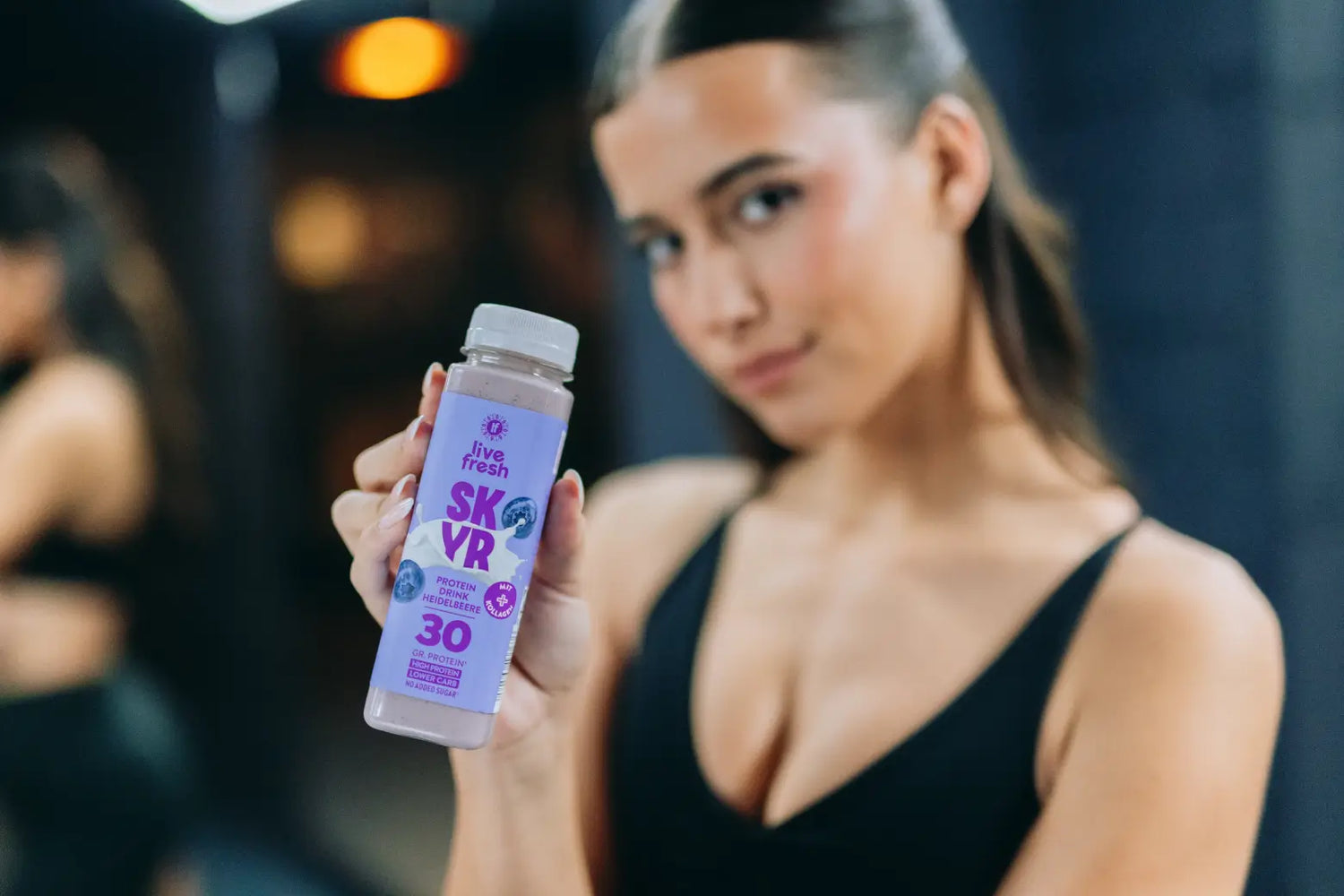 Frau in schwarzer Sportkleidung präsentiert eine Flasche LiveFresh SKYR Protein Drink Heidelbeere mit 30 Gramm Protein im Fitnessstudio.