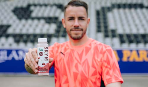 
Ein Fußballspieler im roten Trikot hält einen Skyr Protein Drink mit Kirschgeschmack von Live Fresh in die Kamera, der 30g Protein enthält