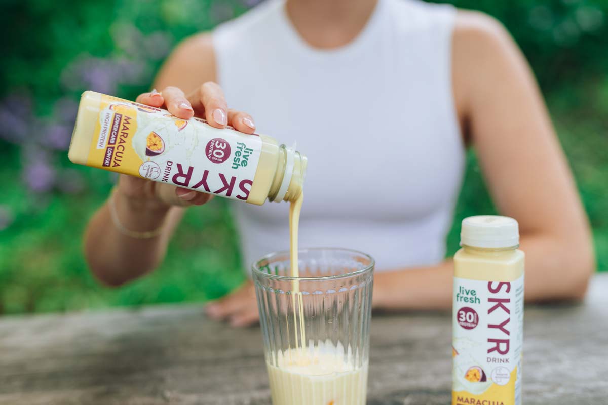 Eine Person gießt einen Live Fresh Skyr Drink mit Maracuja-Geschmack in ein Glas, während eine weitere Flasche Skyr Drink auf dem Tisch steht. Auf der Flasche sind 30g Protein und "Lower Carb, Low Fat" hervorgehoben.