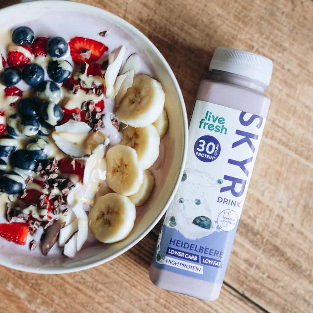 Eine gesunde Bowl, garniert mit Bananenscheiben, Blaubeeren, Erdbeeren, Kokosflocken und Kakaonibs. Daneben steht eine Flasche „Live Fresh Skyr Drink Heidelbeere“ mit 30g Protein. Die Skyr-Flasche wirbt mit einem niedrigen Kohlenhydratgehalt, fettarm und proteinreich, und hebt damit eine ausgewogene, nahrhafte Mahlzeit oder Snack hervor.