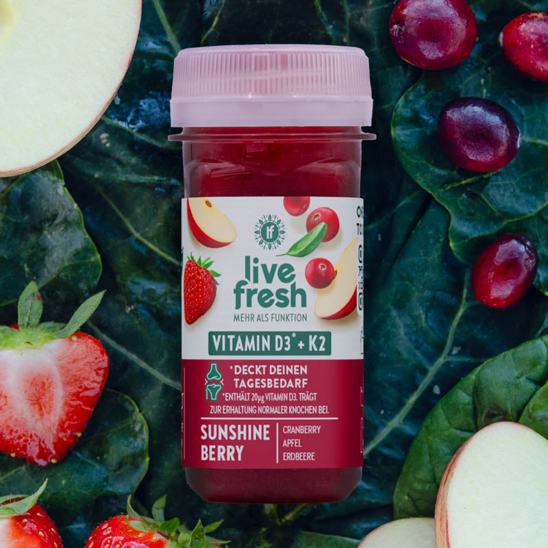 LiveFresh Shot „Sunshine Berry“ mit Vitamin D3 und K2 zur Knochenerhaltung. Kräftig rot, platziert zwischen Apfelstücken, Cranberries und Erdbeeren auf grünem Gemüse.