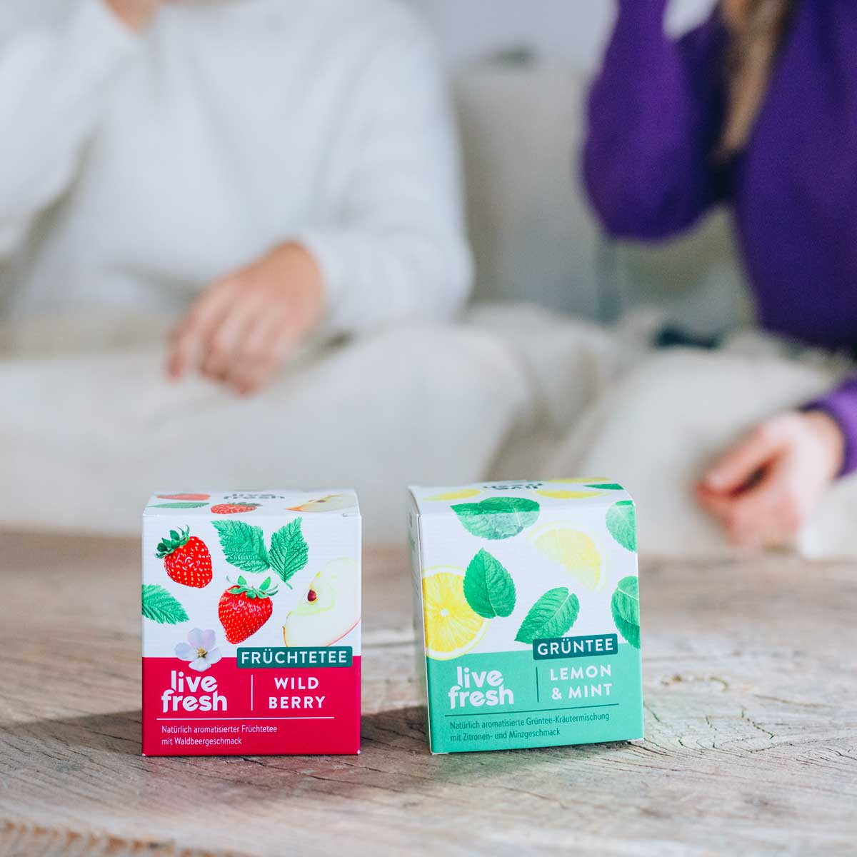 Zwei Packungen LiveFresh Tee auf einem Holztisch, Sorten „Wild Berry“ Früchtetee und „Lemon & Mint“ Grüntee, im Hintergrund sitzen zwei Personen gemütlich auf dem Sofa.
