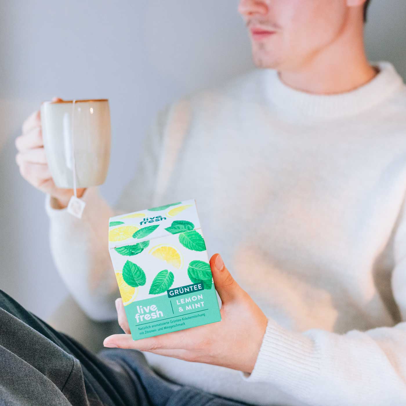 Mann in weißem Pullover hält eine Tasse Tee und eine Packung LiveFresh Grüntee „Lemon & Mint“ – entspannter Moment mit Grüntee.
