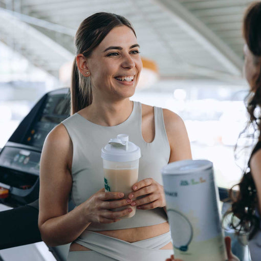 Zwei sportlich gekleidete Frauen unterhalten sich lachend neben dem Laufband im Fitnessstudio, eine hält einen Proteinshake von LiveFresh in  der Hand. 