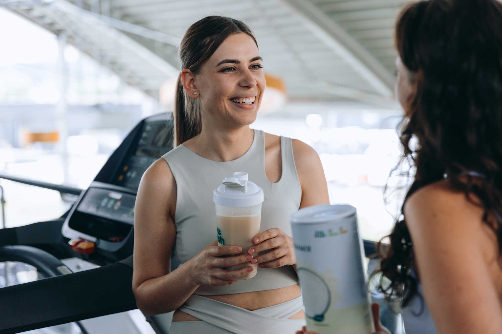 Zwei sportlich gekleidete Frauen unterhalten sich lachend neben dem Laufband im Fitnessstudio, eine hält einen Proteinshake von LiveFresh in  der Hand. 