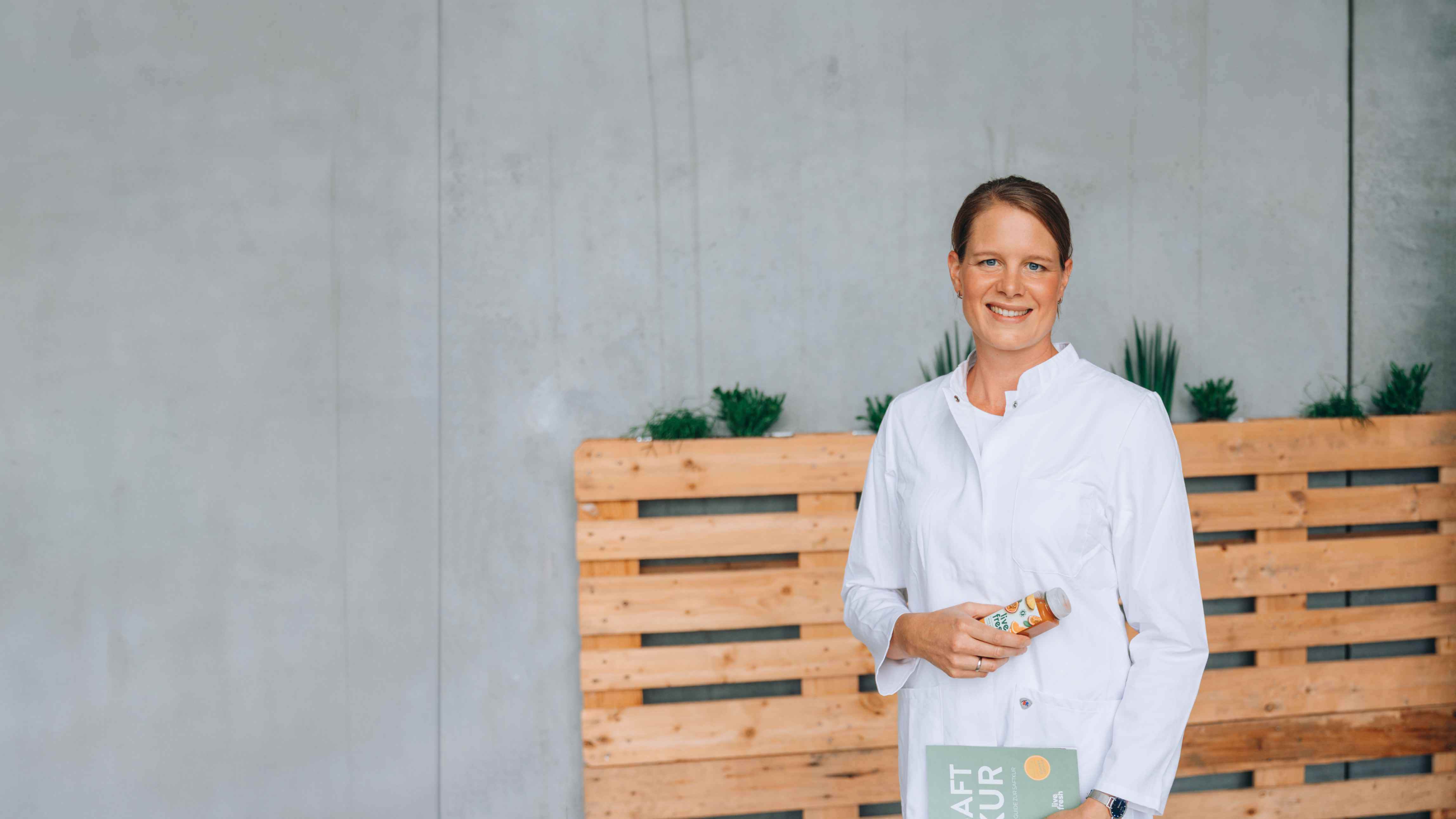Eine lächelnde Frau mit braunen, zusammengebundenen Haaren in einem weißen Kittel hält eine Flasche Saft von LiveFresh in der einen Hand und ein Buch mit der Aufschrift "FOOD KUR" in der anderen Hand. Im Hintergrund stehen Pflanzen auf Holzpaletten vor einer grauen Wand.