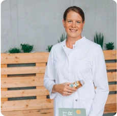 Eine lächelnde Frau mit braunen, zusammengebundenen Haaren in einem weißen Kittel hält eine Flasche Saft von LiveFresh in der einen Hand und ein Buch mit der Aufschrift 