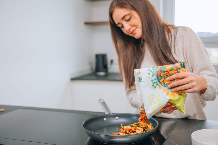 Eine brünette Frau in heller Kleidung schüttet eine LiveFresh Bowl mit buntem Gemüse in eine Pfanne. Sie befindet sich in einer hellen Küche mit dunkler Arbeitsfläche. 