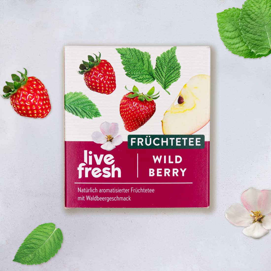 Nahaufnahme der LiveFresh Teeverpackung „Früchtetee Wild Berry“, verziert mit Erdbeeren, Apfelstück, Minzblättern und Blüten auf hellem Hintergrund.