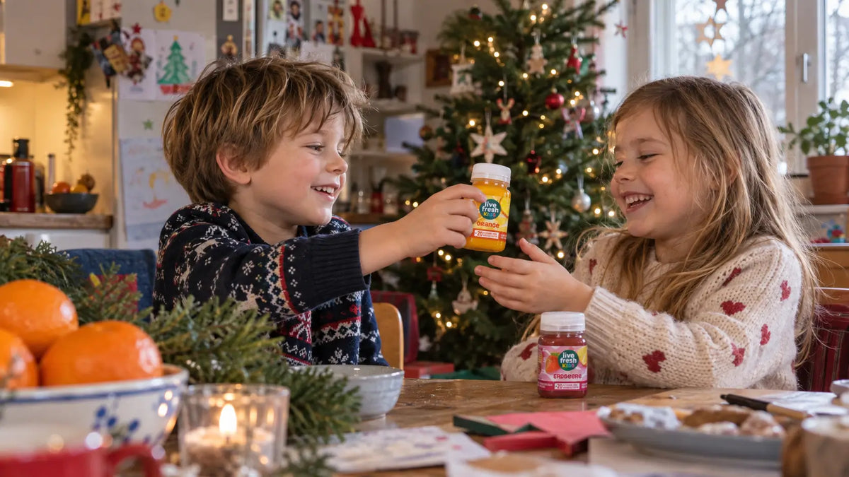 Zwei lachende Kinder sitzen vor einem geschmückten Weihnachtsbaum und reichen sich eine Flasche LiveFresh Kids mit der Aufschrift "ORANGE". Auf dem festlich gedeckten Tisch steht außerdem eine Flasche mit der Aufschrift "ERDBEERE".