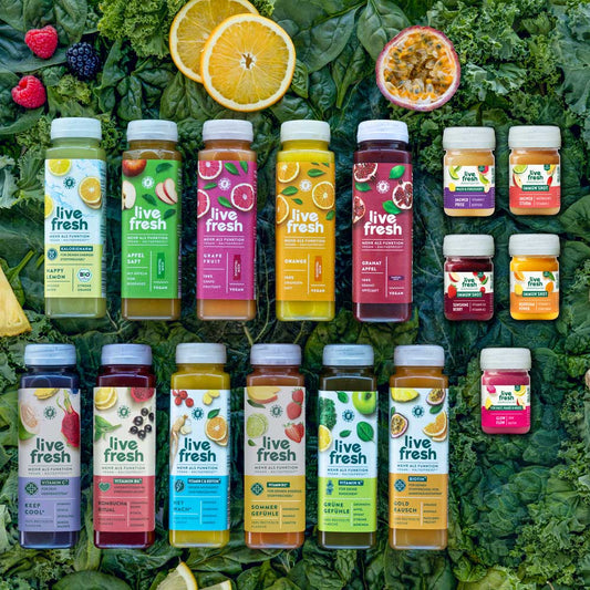 Große Auswahl an LiveFresh Säften und Immun Shots auf einem Hintergrund aus dunklem Blattgemüse mit Beeren Orange Maracuja und Limette. Zu sehen sind zahlreiche Sorten darunter Happy Lemon Apfelsaft Grapefruit Orange Granatapfel Keep Cool Kombucha Ritual Hey Wach Sommer Gefühle Grüne Gefühle Gold Rausch sowie verschiedene Immun Shots.