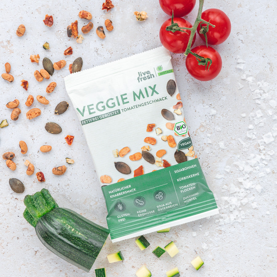 Packung von 'Live Fresh Veggie Mix' mit Tomatengeschmack und Streuung von Zucchini und Tomaten neben der Packung auf einer hellen Oberfläche, hervorragend als glutenfreier Snack.